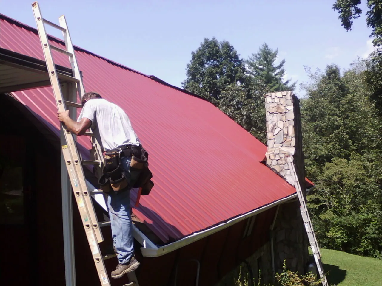 Roofer on a ladder beside a red metal roof and stone chimney — Soffit & Fascia Repair by Roofing Brothers in Reform